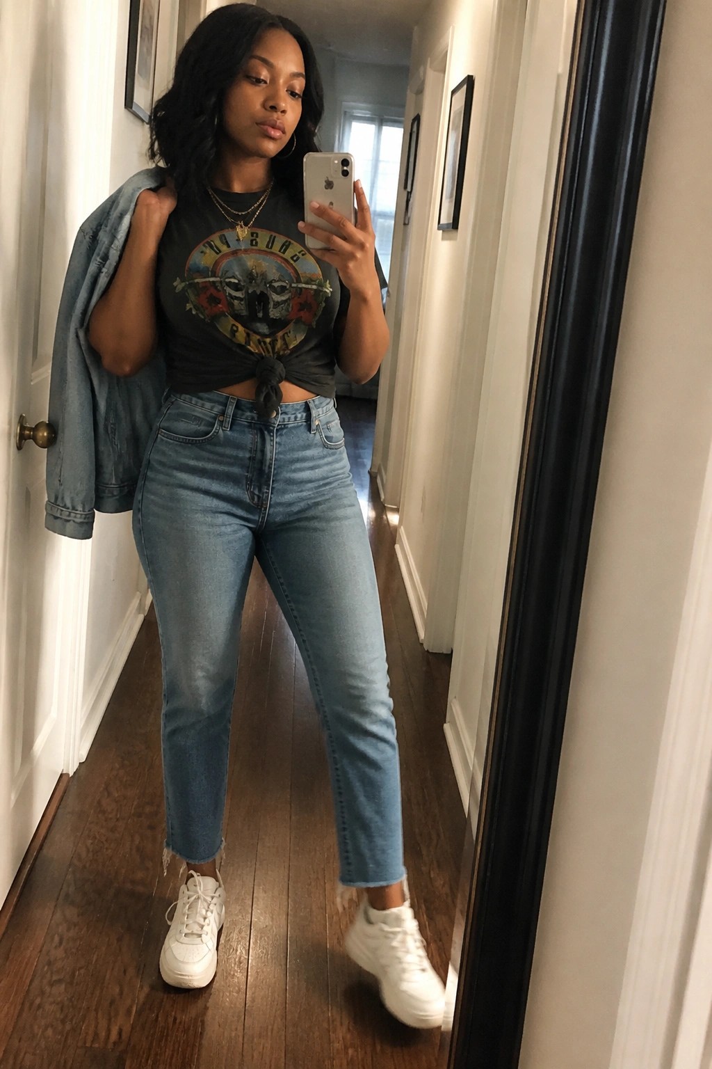 Woman in knotted black graphic tee and jeans takes mirror selfie in hallway.