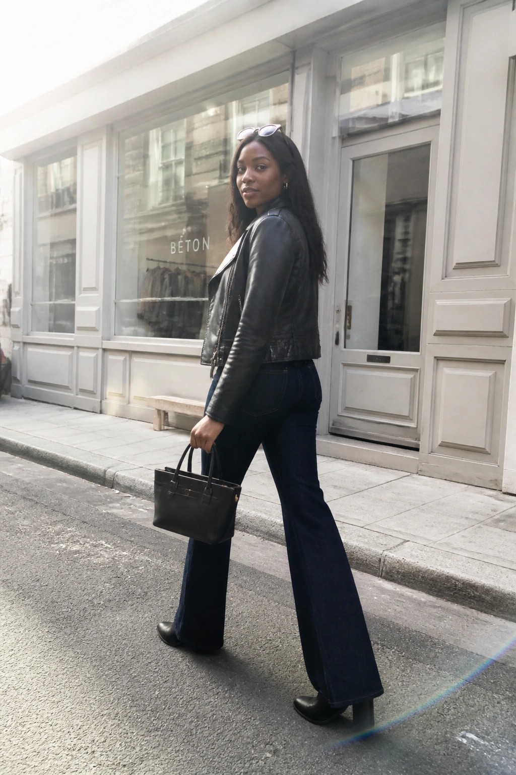 Woman in black leather jacket and flared jeans holding handbag on sidewalk