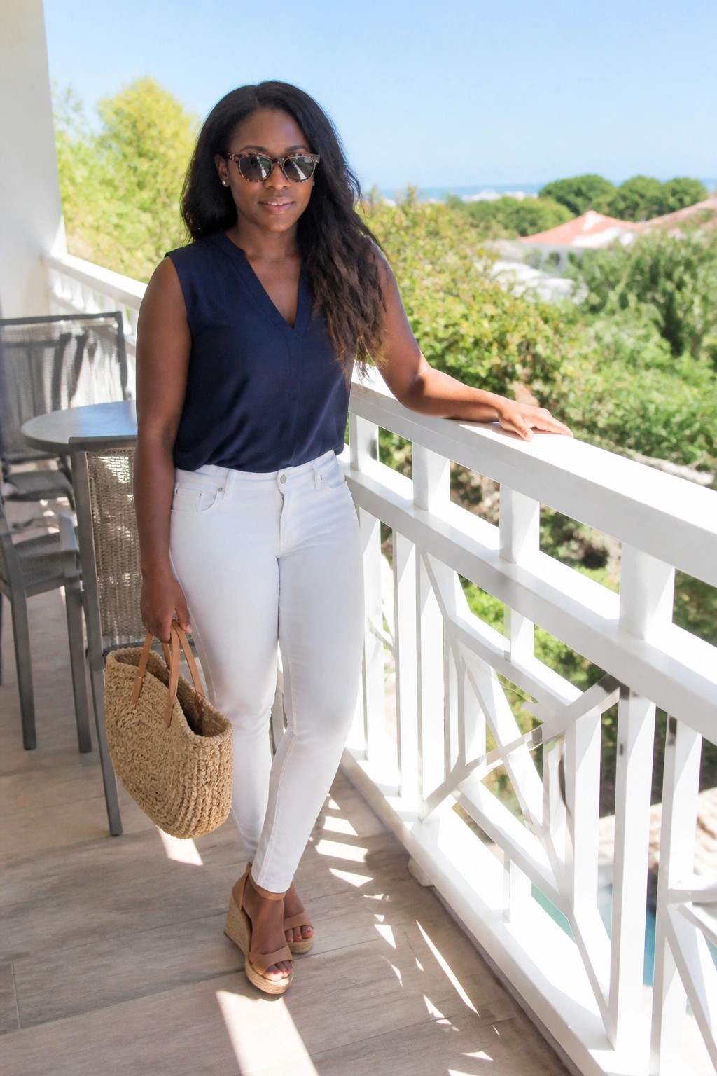 Woman wearing a navy sleeveless V-neck top tucked into white skinny jeans with brown wedge sandals and carrying a straw tote bag.