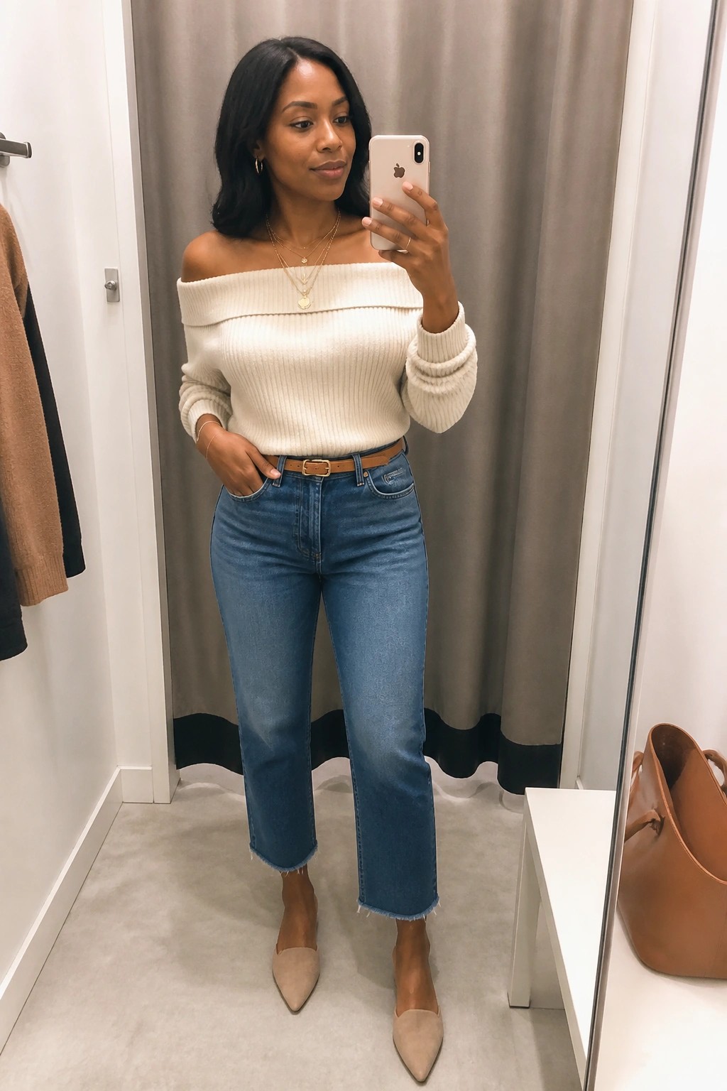 A Black woman taking a mirror selfie in a fitting room wearing an off-the-shoulder cream ribbed sweater, blue straight-leg jeans with a brown belt, beige pointed flats, and layered gold necklaces.