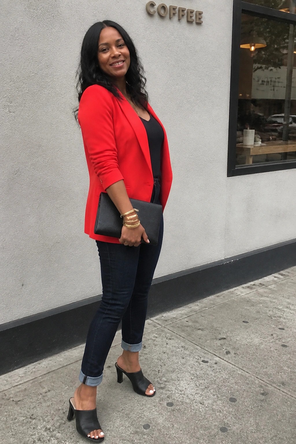 Woman in red blazer and jeans holding black clutch outside coffee shop