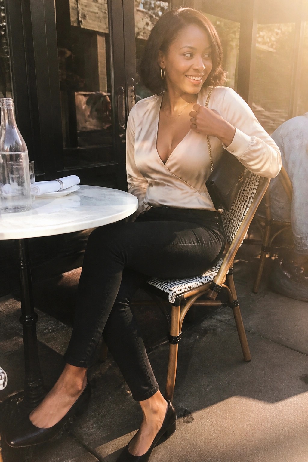 Woman in beige satin blouse and black jeans at sunny outdoor cafe table