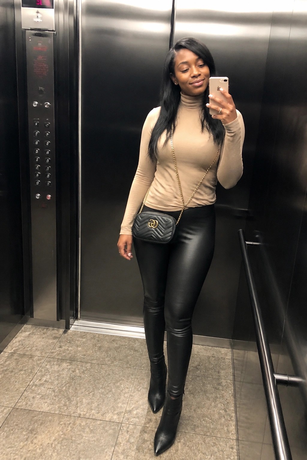 Woman in beige turtleneck and black leather pants taking elevator selfie with handbag