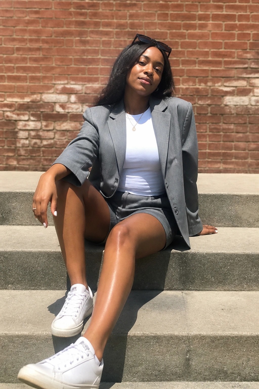 Woman in gray blazer, white top, and shorts sitting on concrete steps by brick wall.