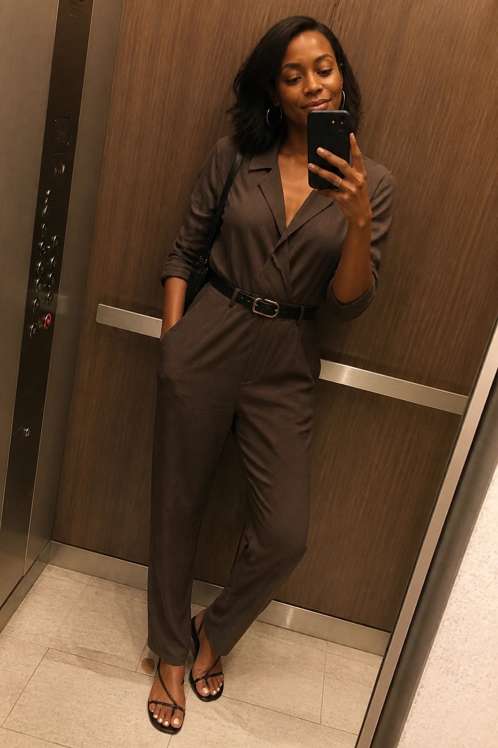 Woman in brown belted jumpsuit taking selfie in wood-paneled elevator