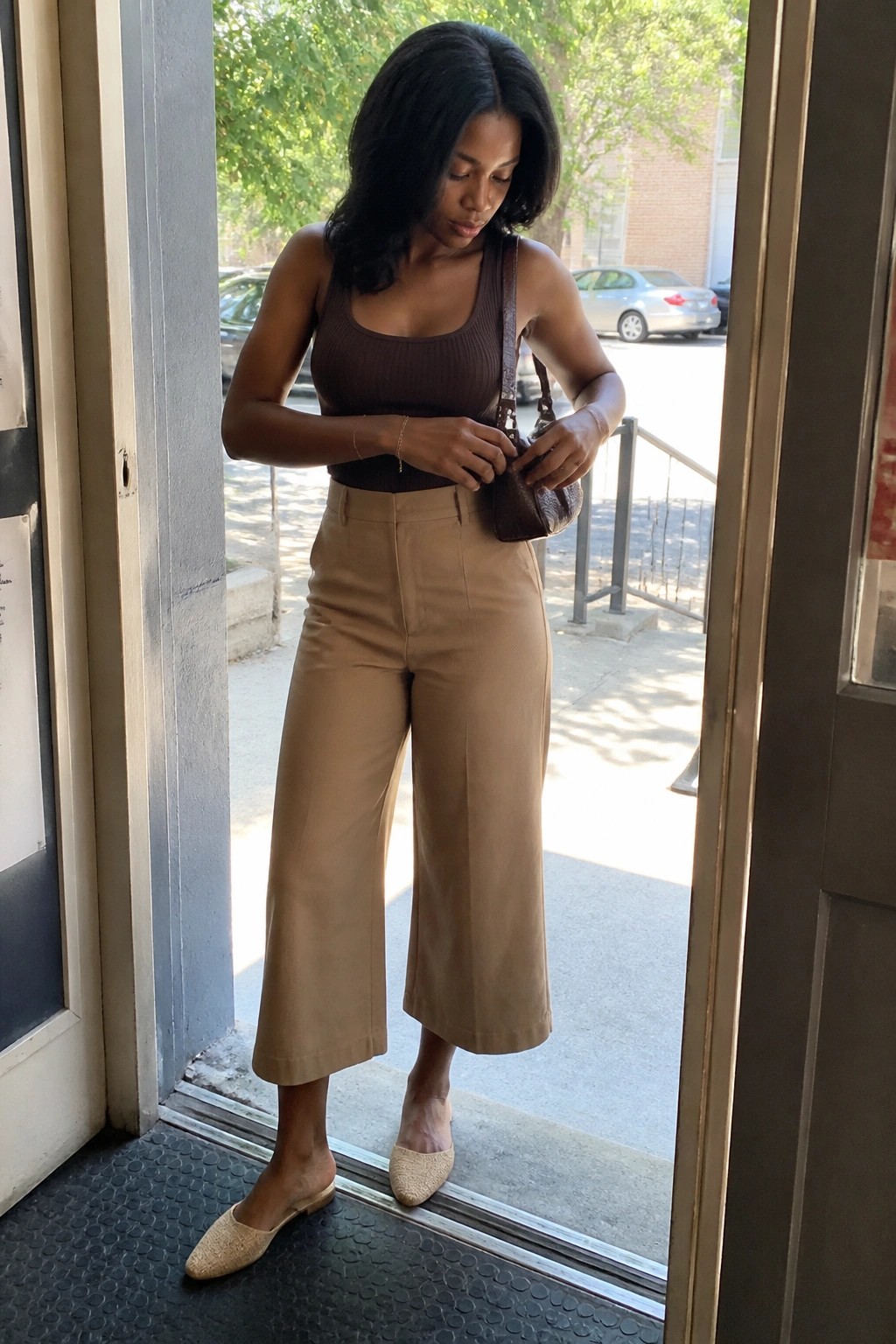Woman in brown tank top and tan wide-leg pants adjusts brown purse in doorway