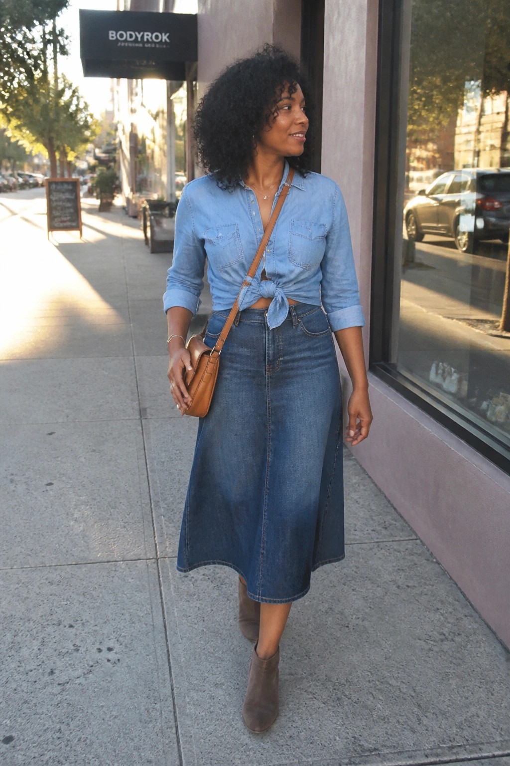 Woman in tied light-blue denim shirt and midi skirt with brown bag on sidewalk
