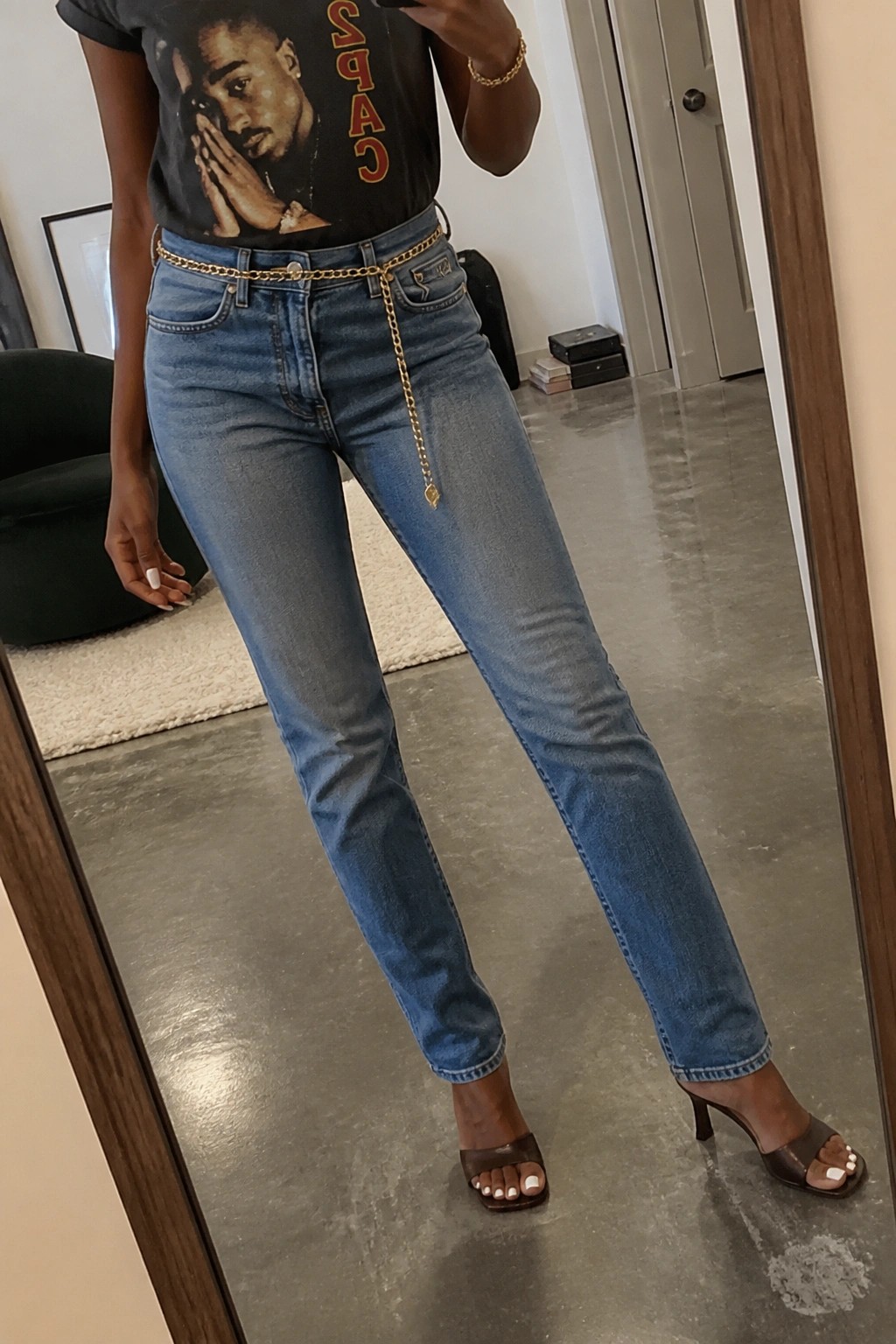Woman in black Tupac t-shirt, blue jeans with gold chain belt, brown heels.