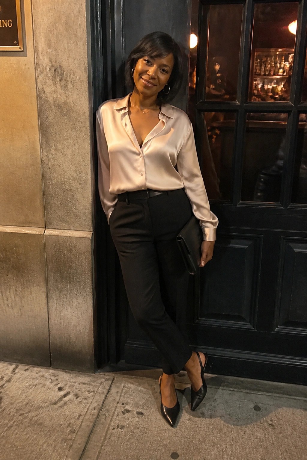 Woman in beige satin blouse and black trousers holding clutch by doorway