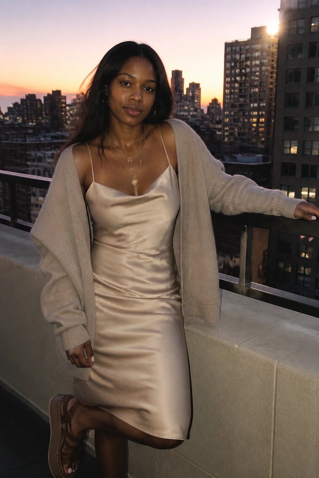 Woman in beige satin slip dress and fuzzy cardigan on city balcony at sunset
