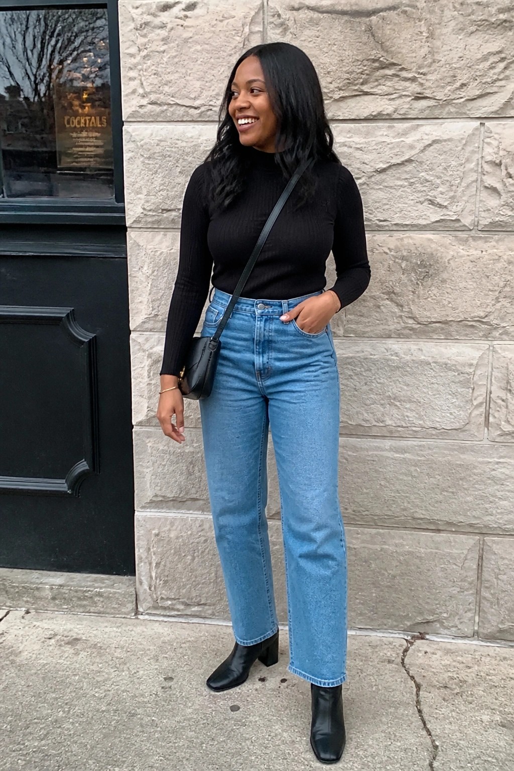 Smiling woman in black turtleneck, blue jeans, and black boots by stone wall
