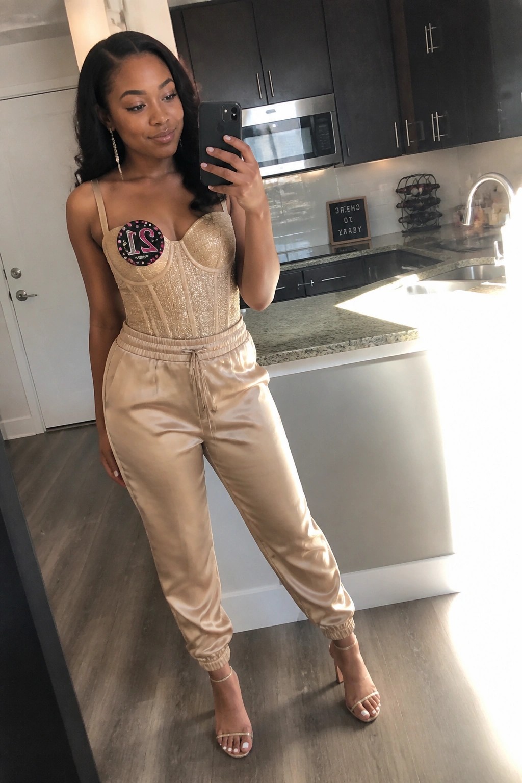 Woman in sparkly gold corset and beige satin joggers taking kitchen selfie