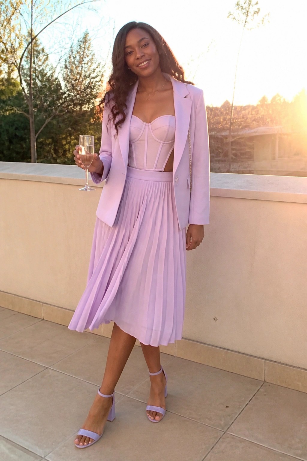Woman in lavender blazer, corset top, and pleated skirt holding champagne at sunset.