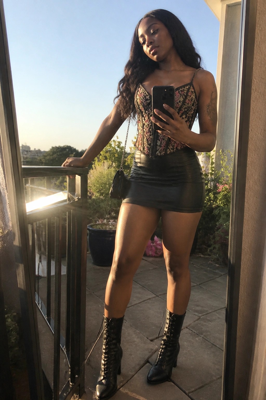 Woman in floral embroidered corset top black leather mini skirt and black lace up boots holding phone for mirror selfie.