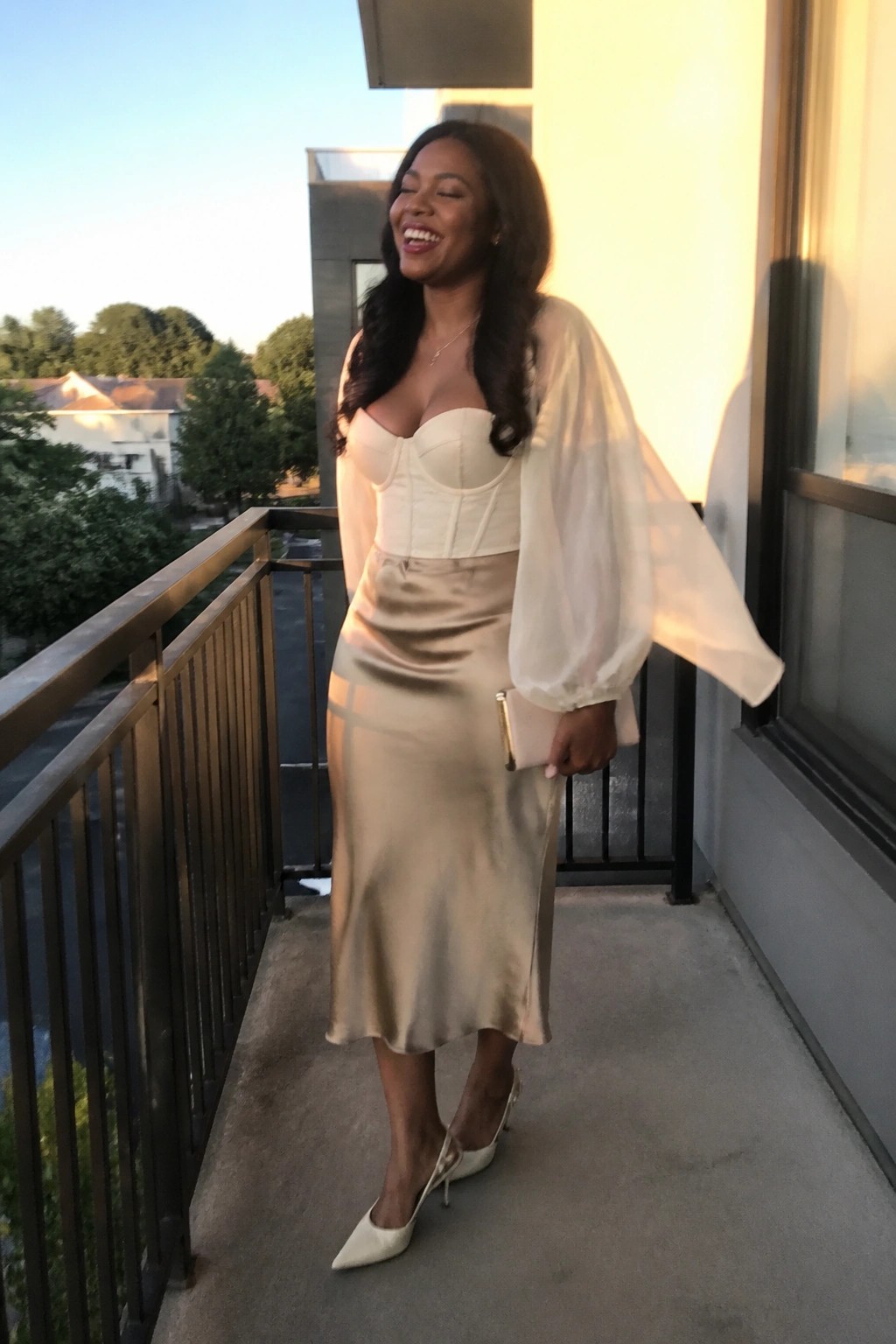 Woman in cream corset with sheer balloon sleeves tucked into a beige satin midi skirt, white pointed heels, and small clutch held at her side.