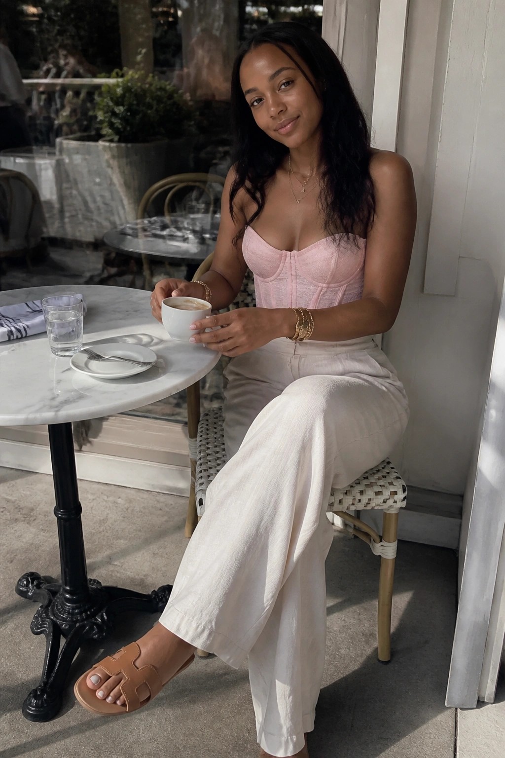 Woman wearing pink strapless corset top with white wide leg pants, gold layered necklaces and bracelets, plus brown open toe sandals while seated at a cafe table holding a white coffee cup.