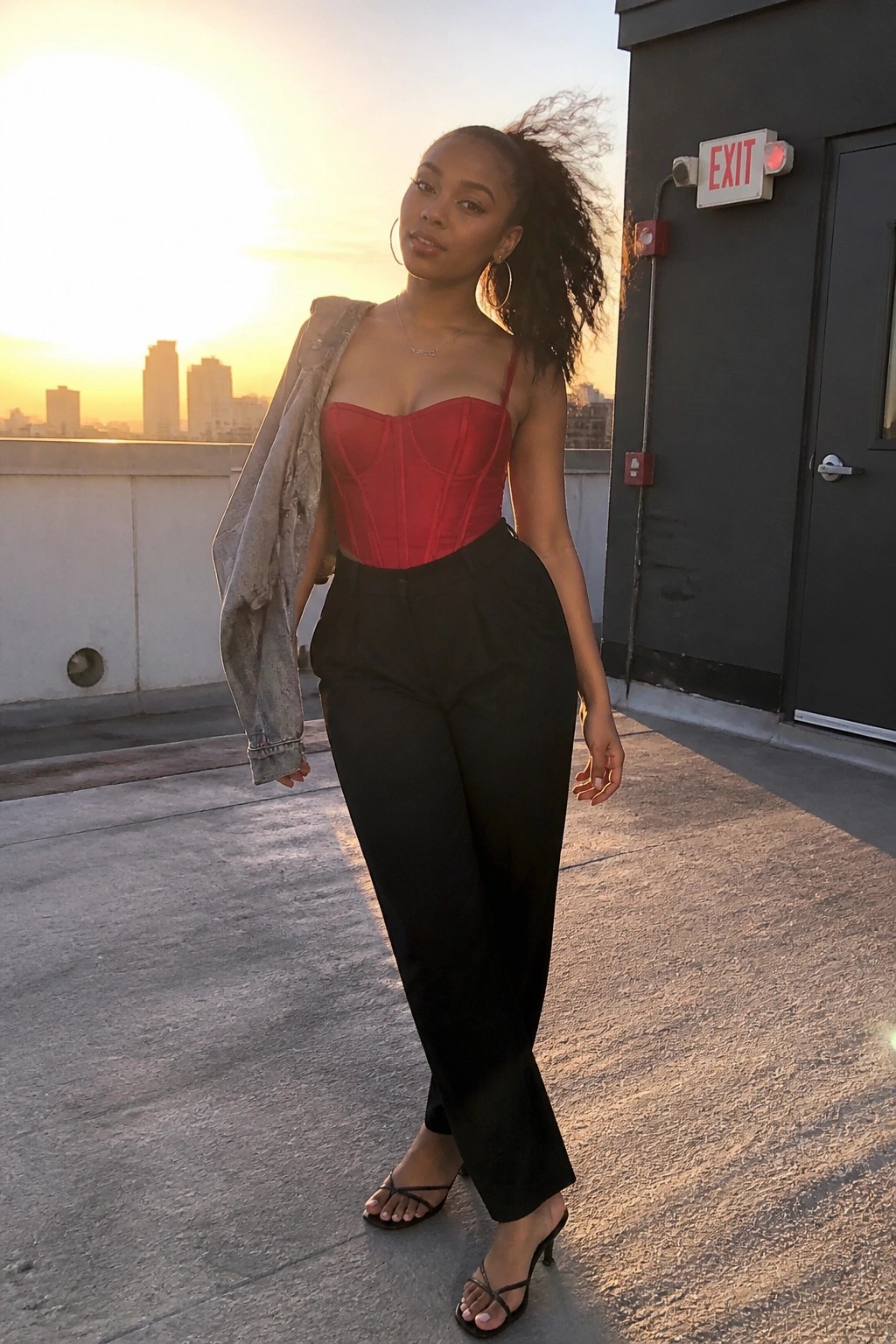 Woman in red corset top and black pants on sunset city rooftop holding denim jacket