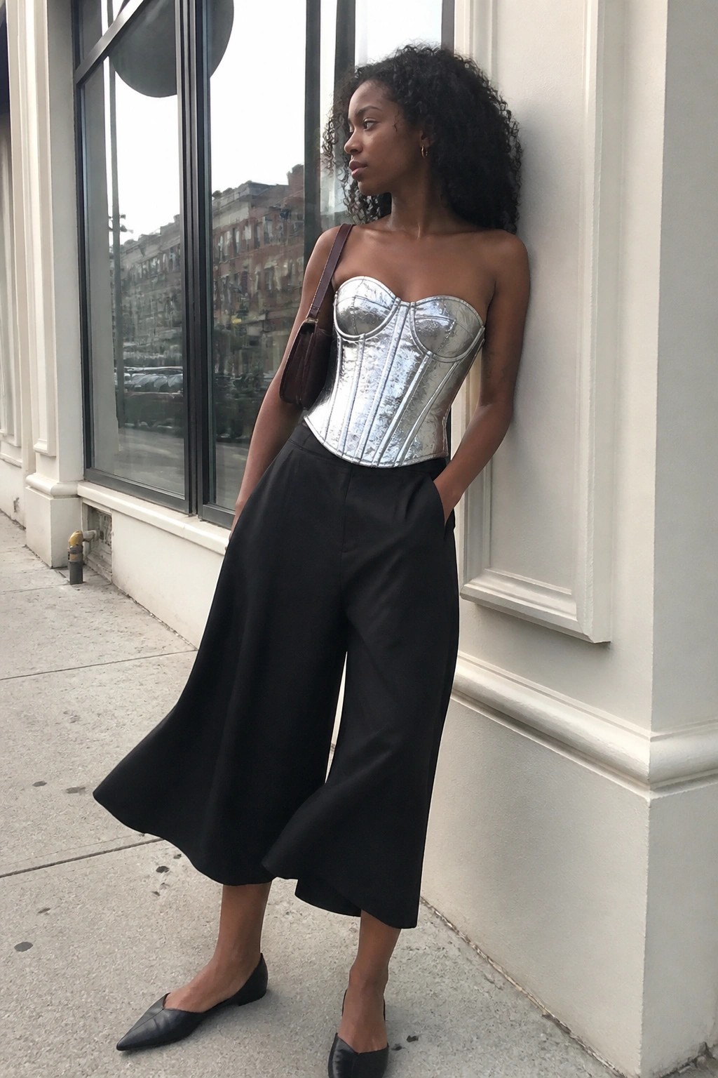 Woman in metallic silver corset and black wide-leg pants leaning on building wall