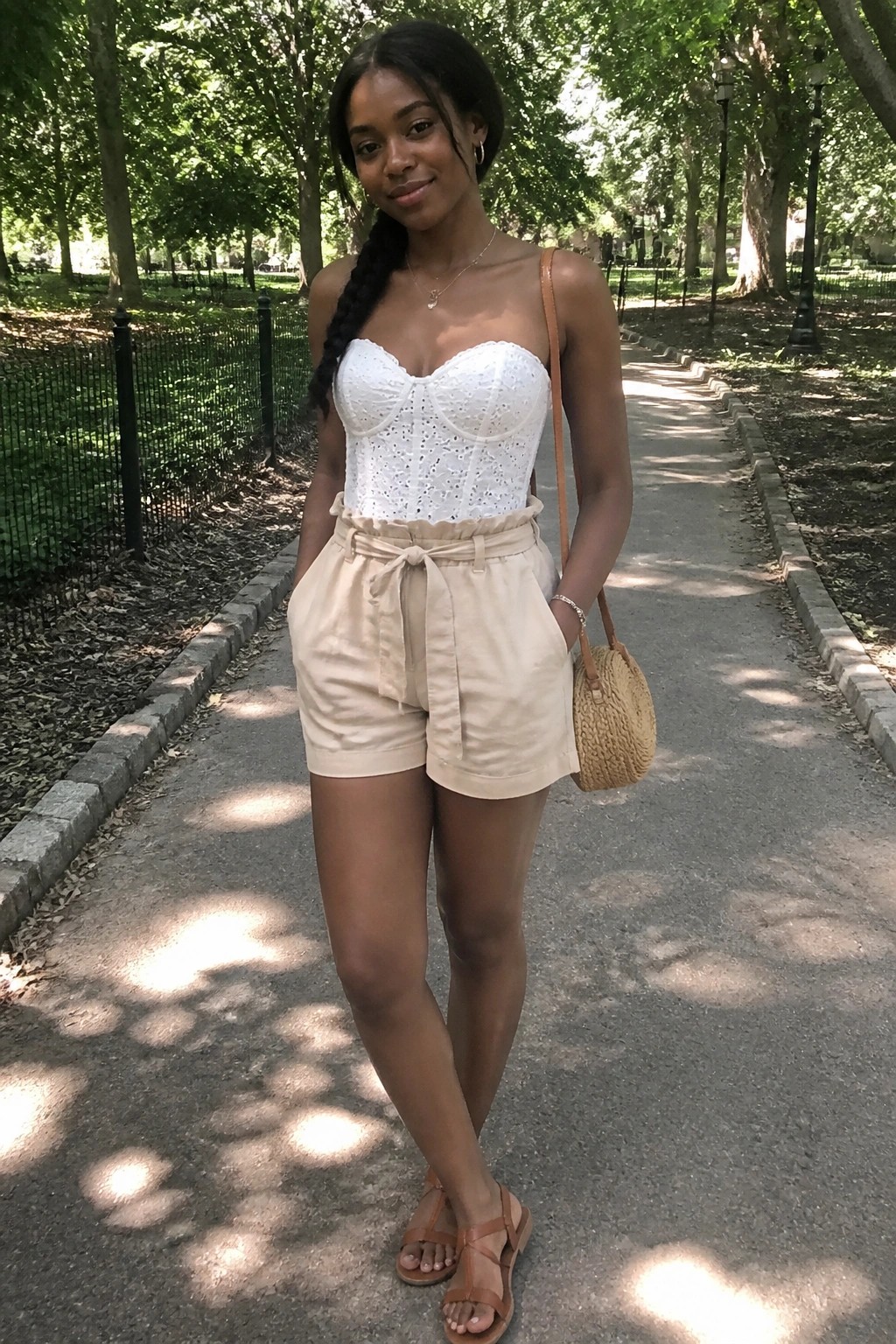 Woman wearing white eyelet corset top and beige paperbag waist shorts with brown sandals and woven bag in a park setting.