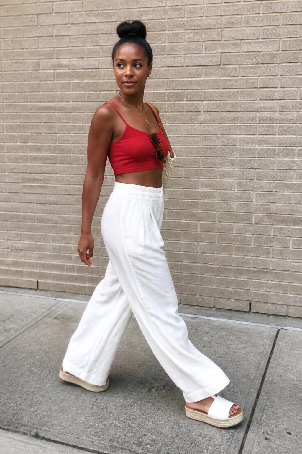 Woman in red crop top and white wide-leg pants walking by brick wall.