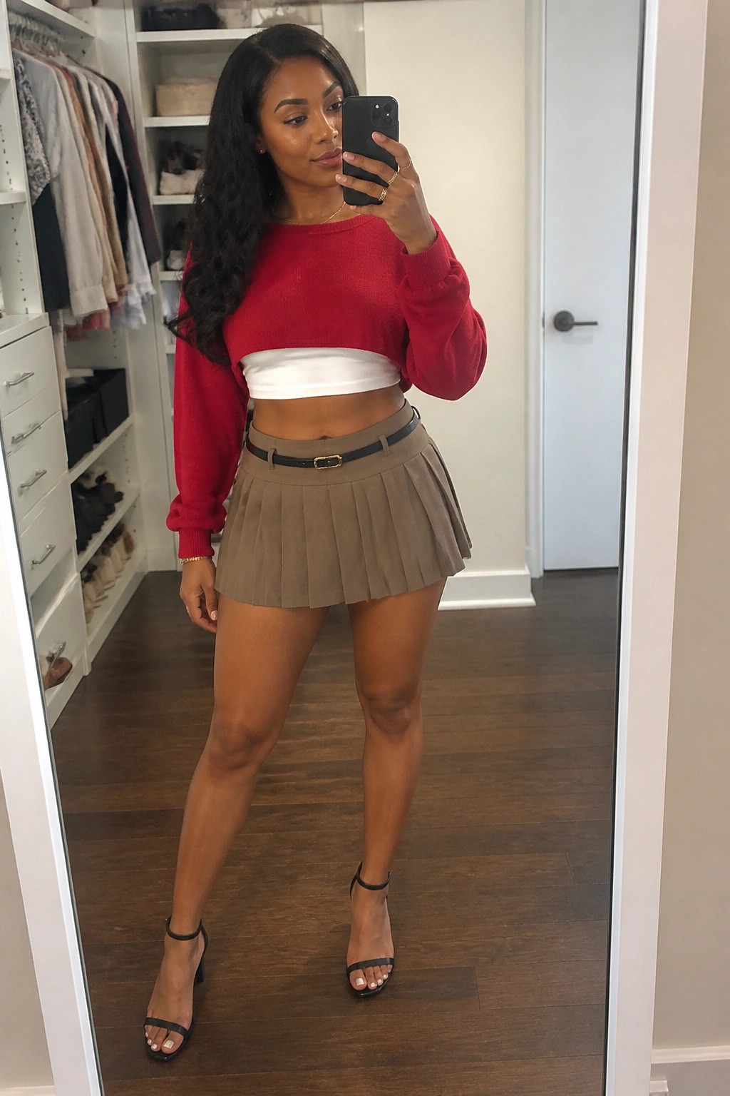 Black woman taking a mirror selfie wearing a red cropped knit sweater over a white bandeau top, paired with a taupe pleated mini skirt, black belt, and black strappy heels.