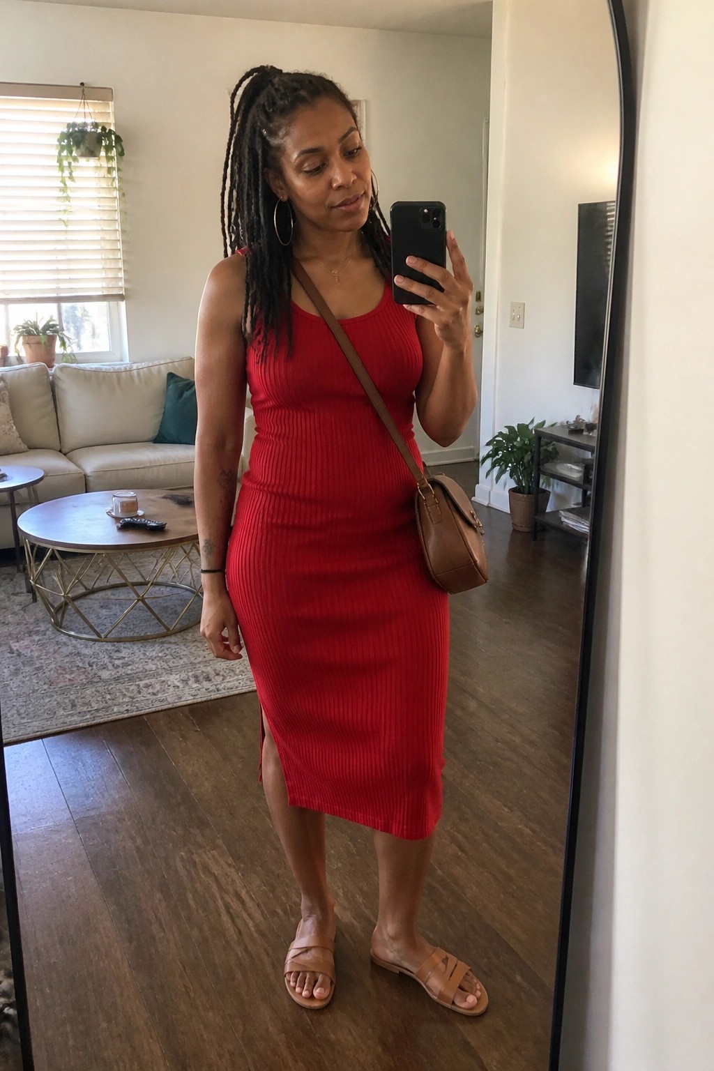 Woman wearing a fitted red ribbed midi dress with a side slit, brown leather crossbody bag, matching brown slide sandals, gold hoop earrings, and thin necklace while taking a mirror selfie in a living room.