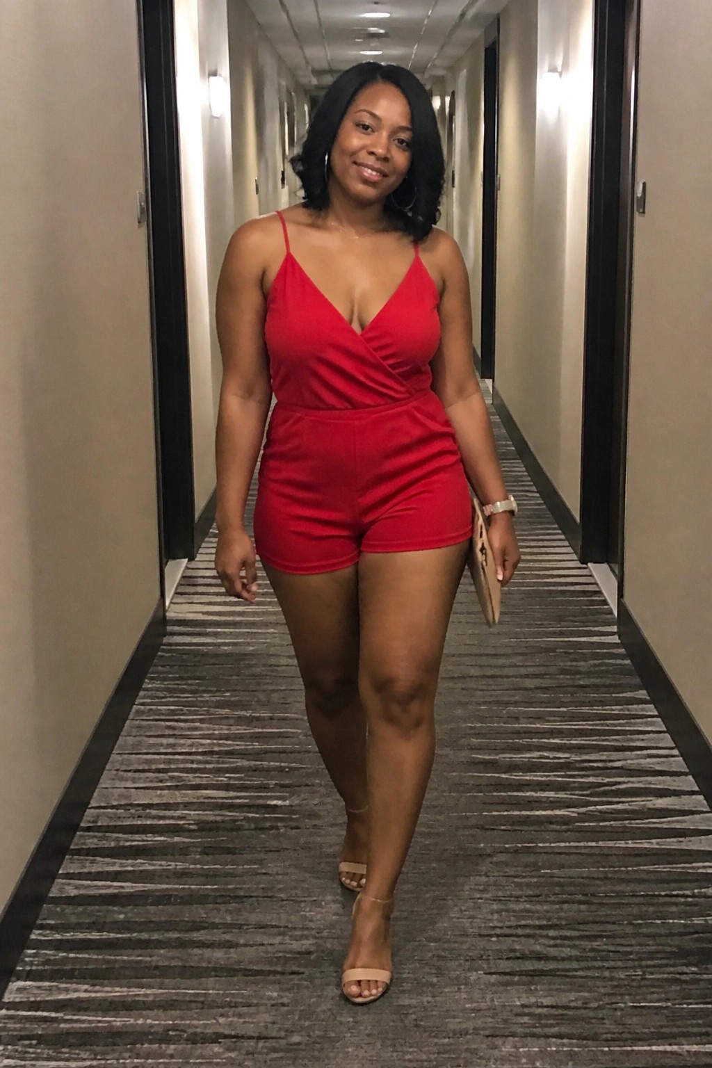 Woman in red romper holding clutch walks down patterned hotel hallway