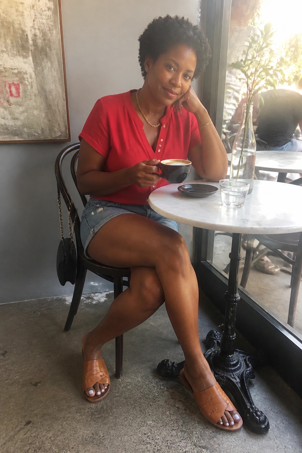 Woman in red top and denim shorts holds coffee at marble cafe table.