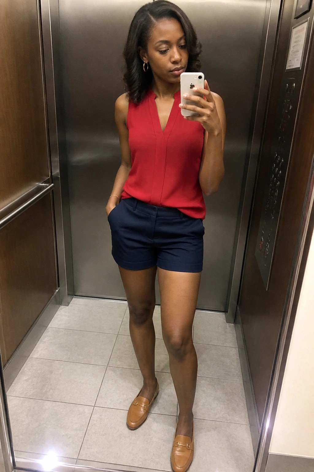 Woman in red top and navy shorts taking selfie in elevator with white phone.