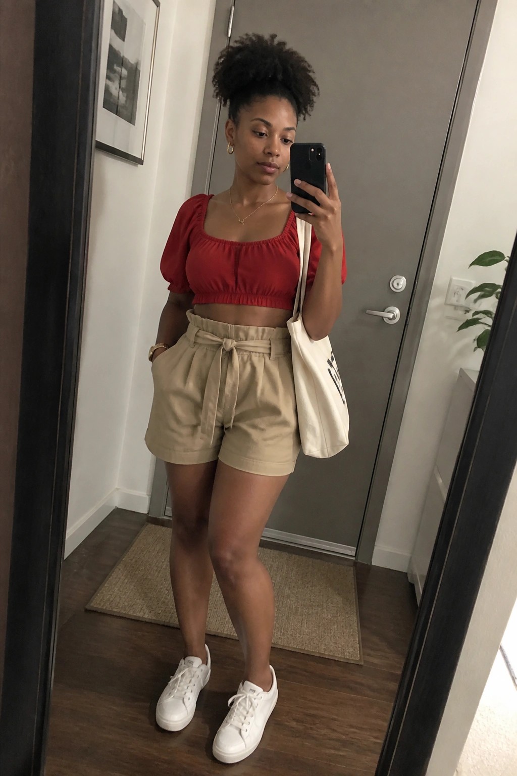 Woman in red crop top and beige shorts taking mirror selfie with tote bag