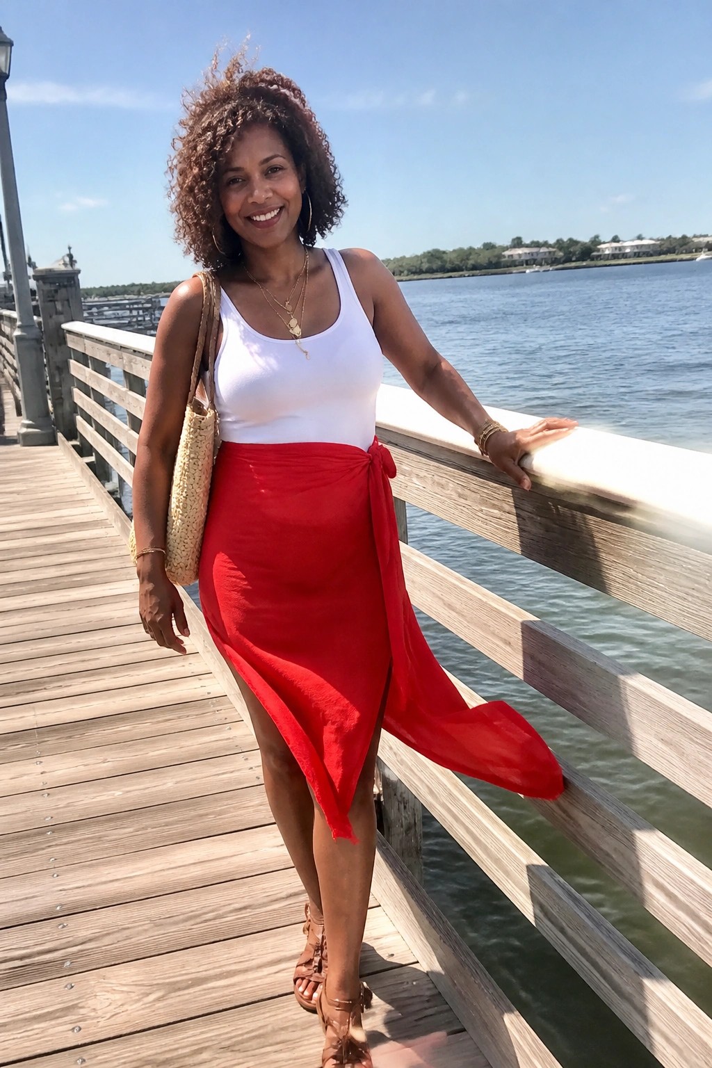 Smiling woman with curly hair in white tank top and red wrap skirt on wooden pier by water