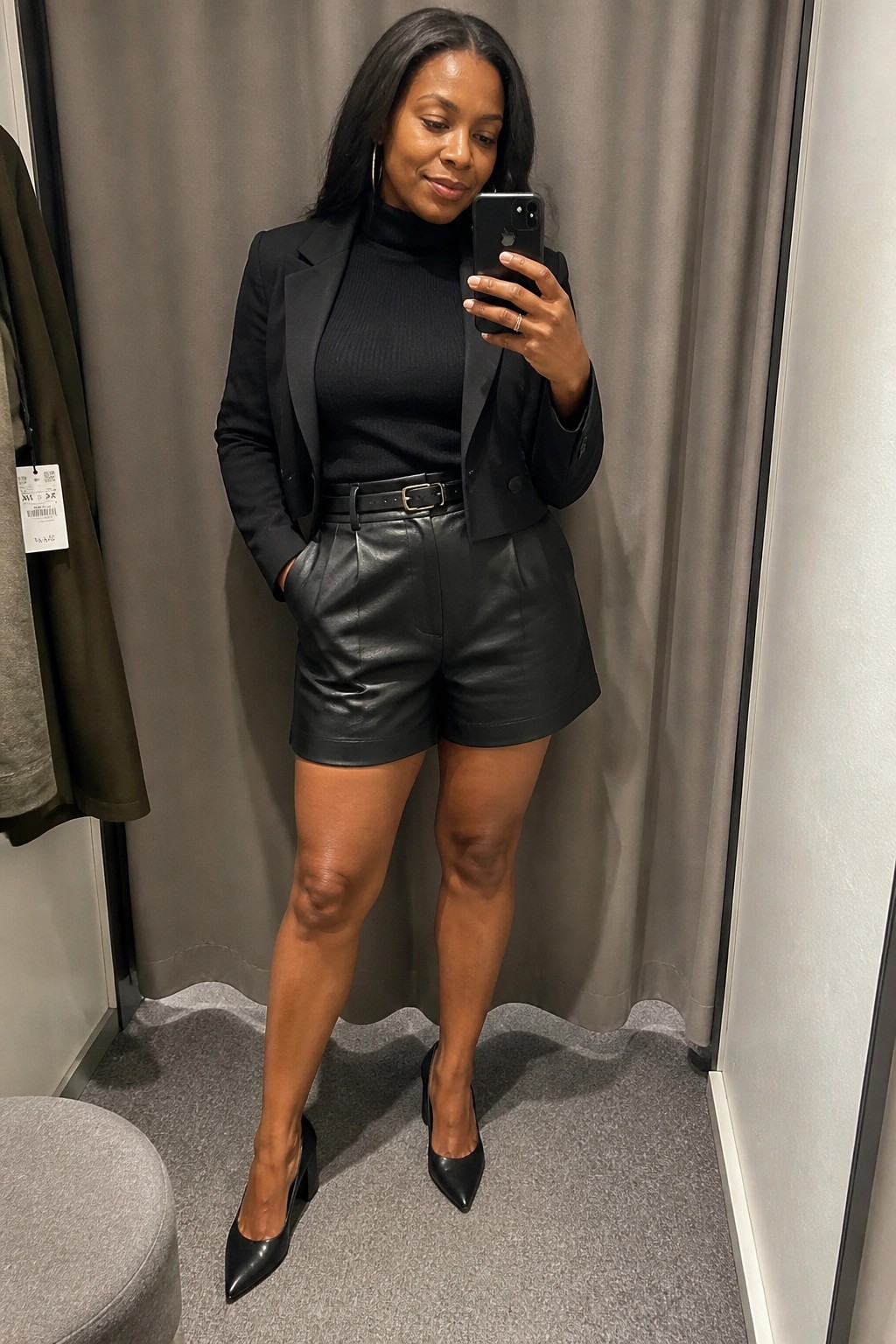 Woman in black turtleneck, blazer, and leather shorts taking mirror selfie in fitting room