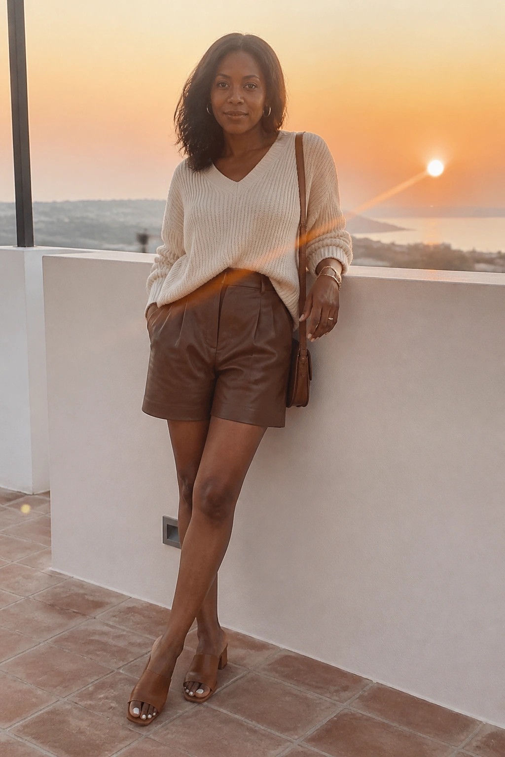 Woman in cream sweater and brown leather shorts at ocean sunset balcony