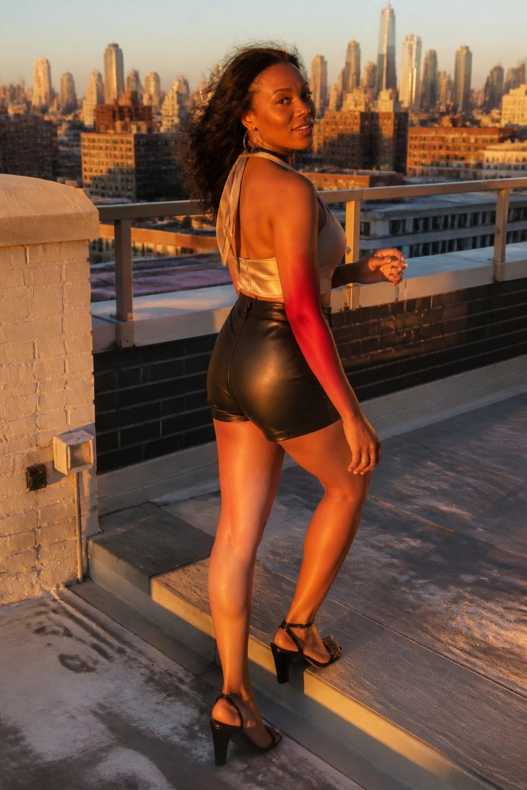 Woman in black leather shorts and beige satin halter top with black strappy heels standing on steps