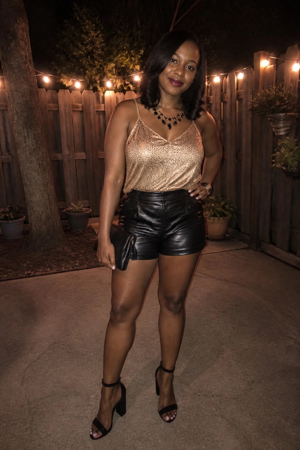 Woman in gold sequined top and black leather shorts at night with string lights.