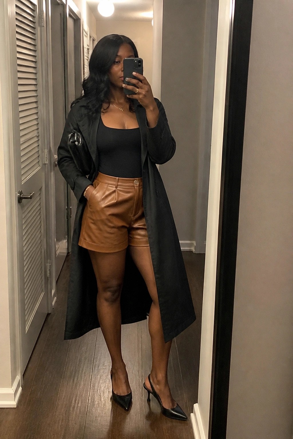 Woman in long black coat and brown leather shorts taking mirror selfie in hallway