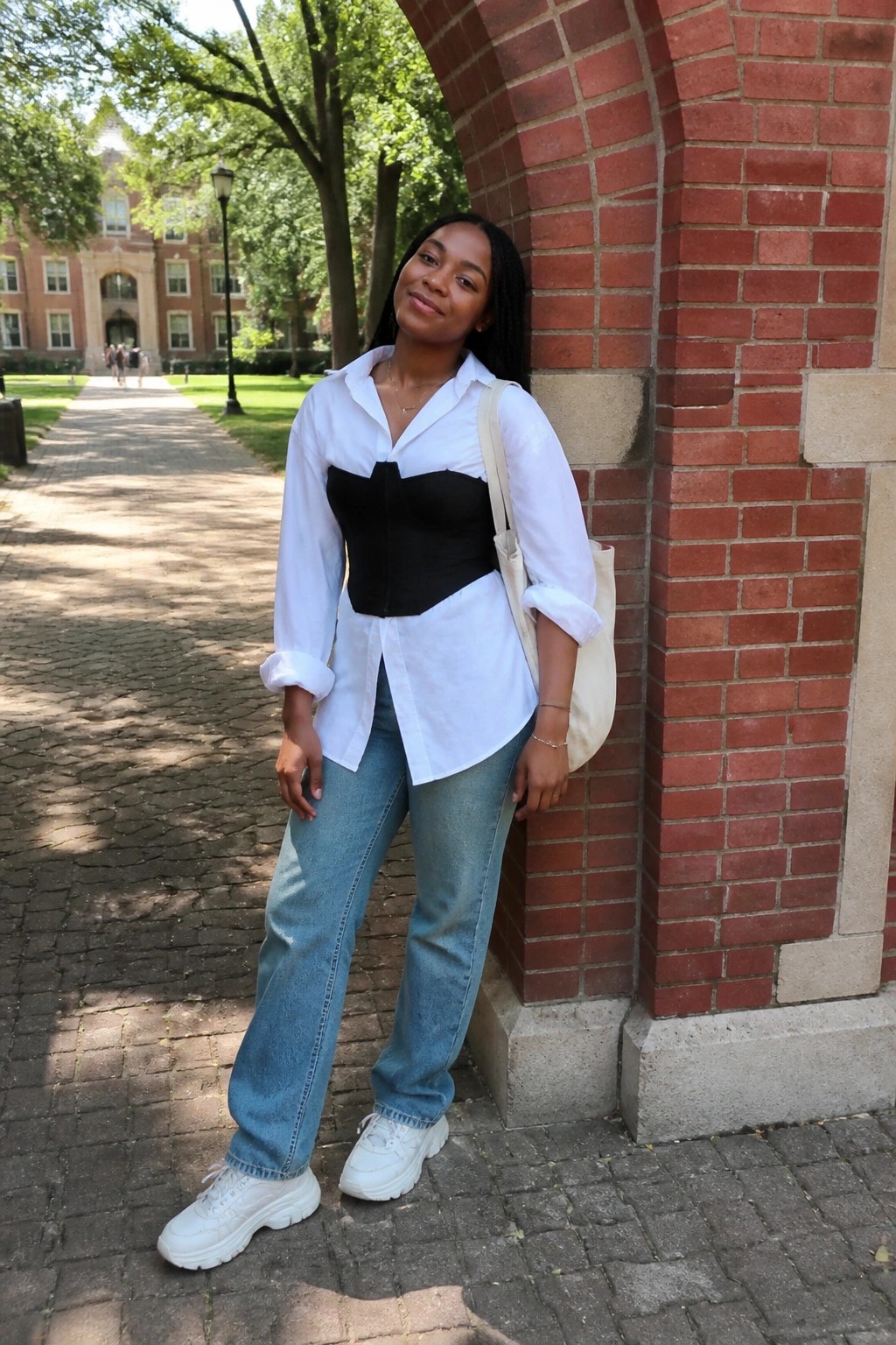 Woman in white shirt over black corset, jeans, and sneakers leaning on brick archway