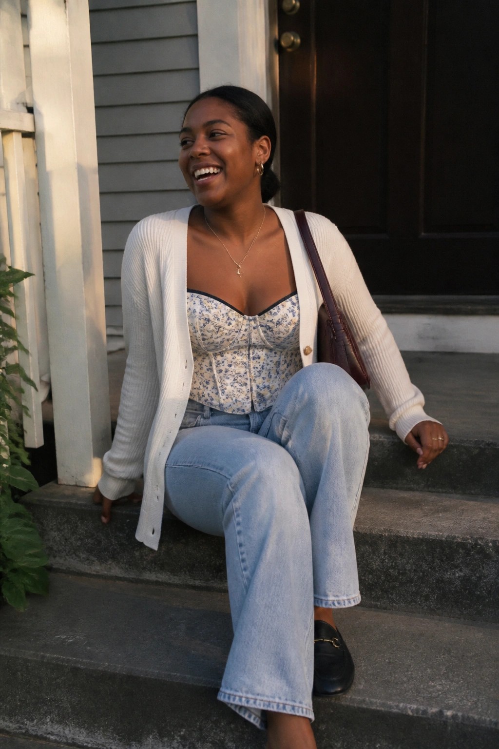 Woman smiles on stone steps in floral corset, white cardigan, and blue jeans
