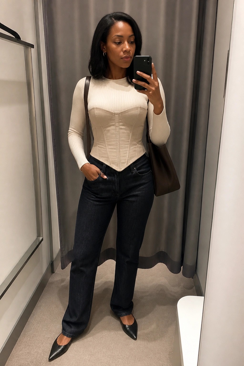 Woman in beige corset and dark jeans taking mirror selfie in fitting room