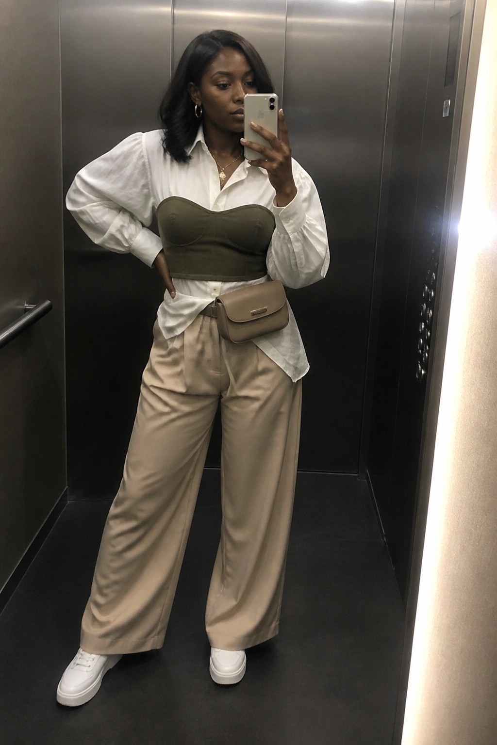 A black woman in an elevator mirror selfie wearing a white oversized button down shirt over an olive green corset top with beige wide leg trousers white sneakers and a beige belt bag.