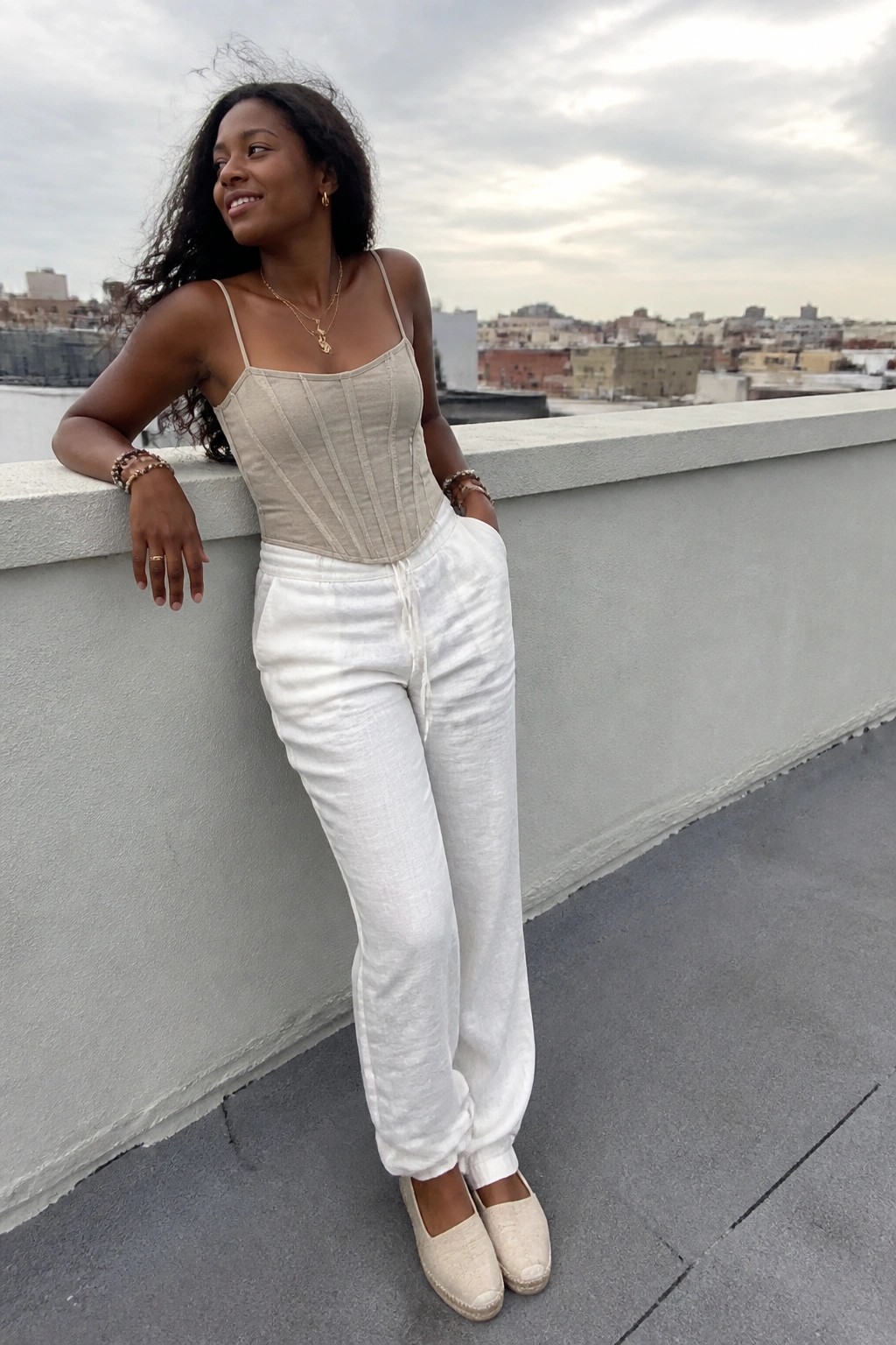 Woman in beige corset top and white pants leaning on rooftop ledge overlooking city