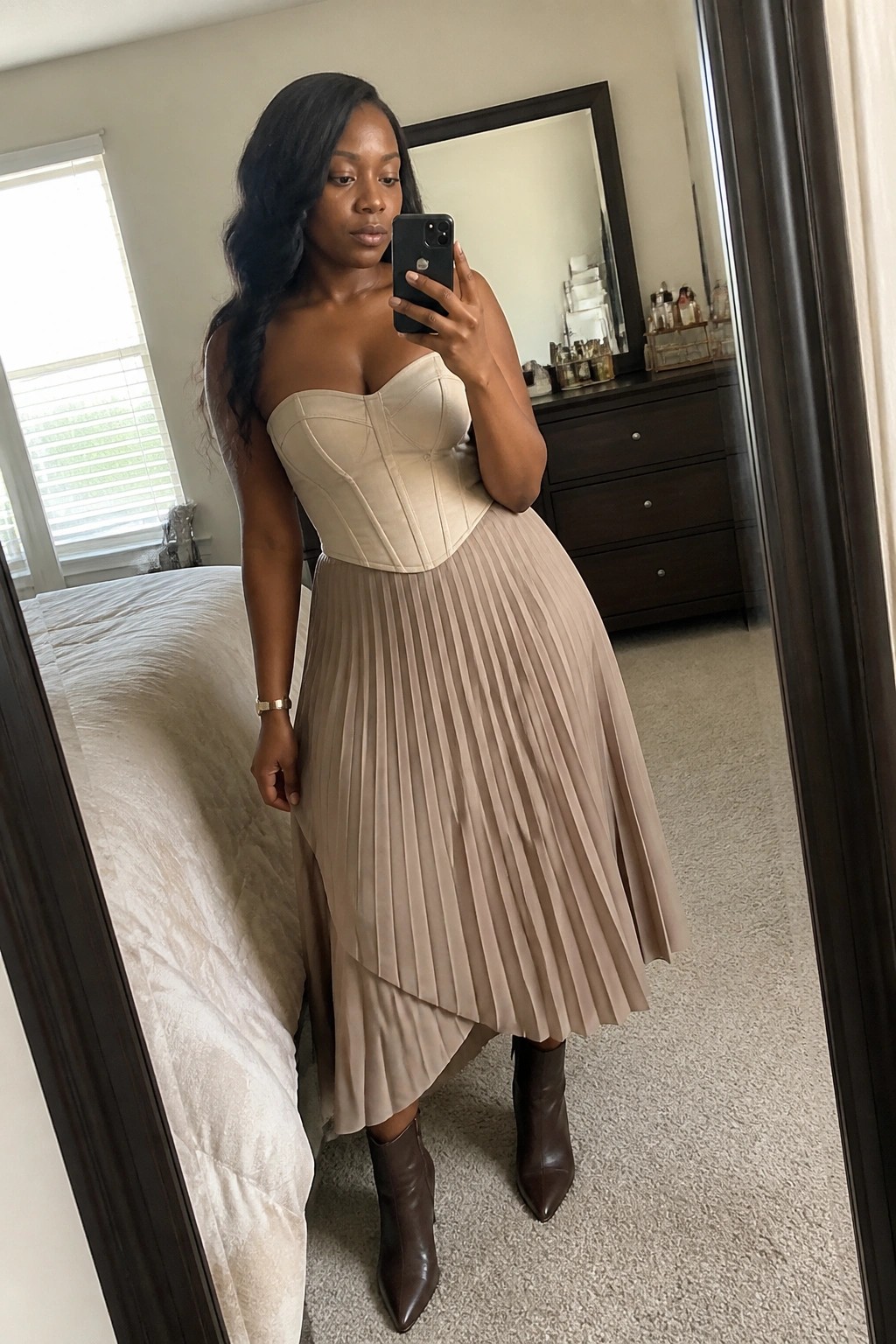 Woman in beige corset top and pleated skirt taking mirror selfie in bedroom