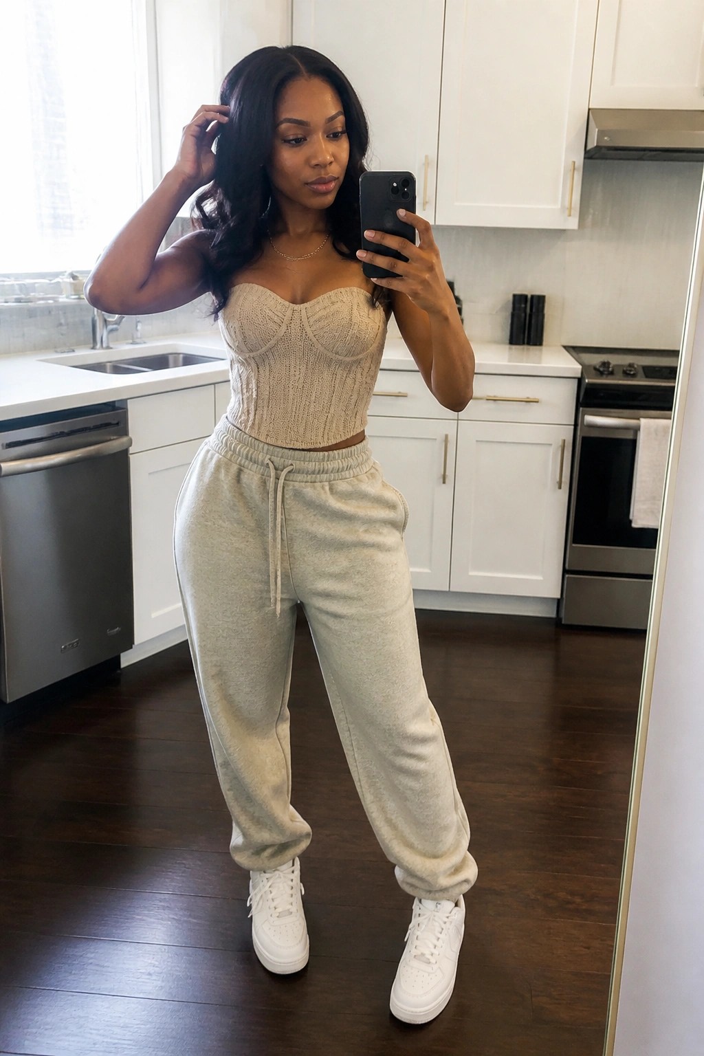 Woman in beige knit corset top, light gray sweatpants, and white sneakers taking a mirror selfie in a kitchen setting.