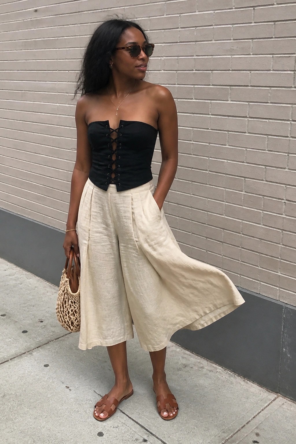 A Black woman in a black strapless lace-up corset top paired with cream wide-leg linen pants, brown flat sandals, and a woven straw bag, standing on a sidewalk.