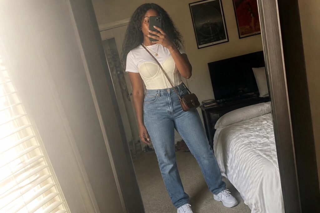 Woman in beige corset over white tee with blue jeans taking mirror selfie