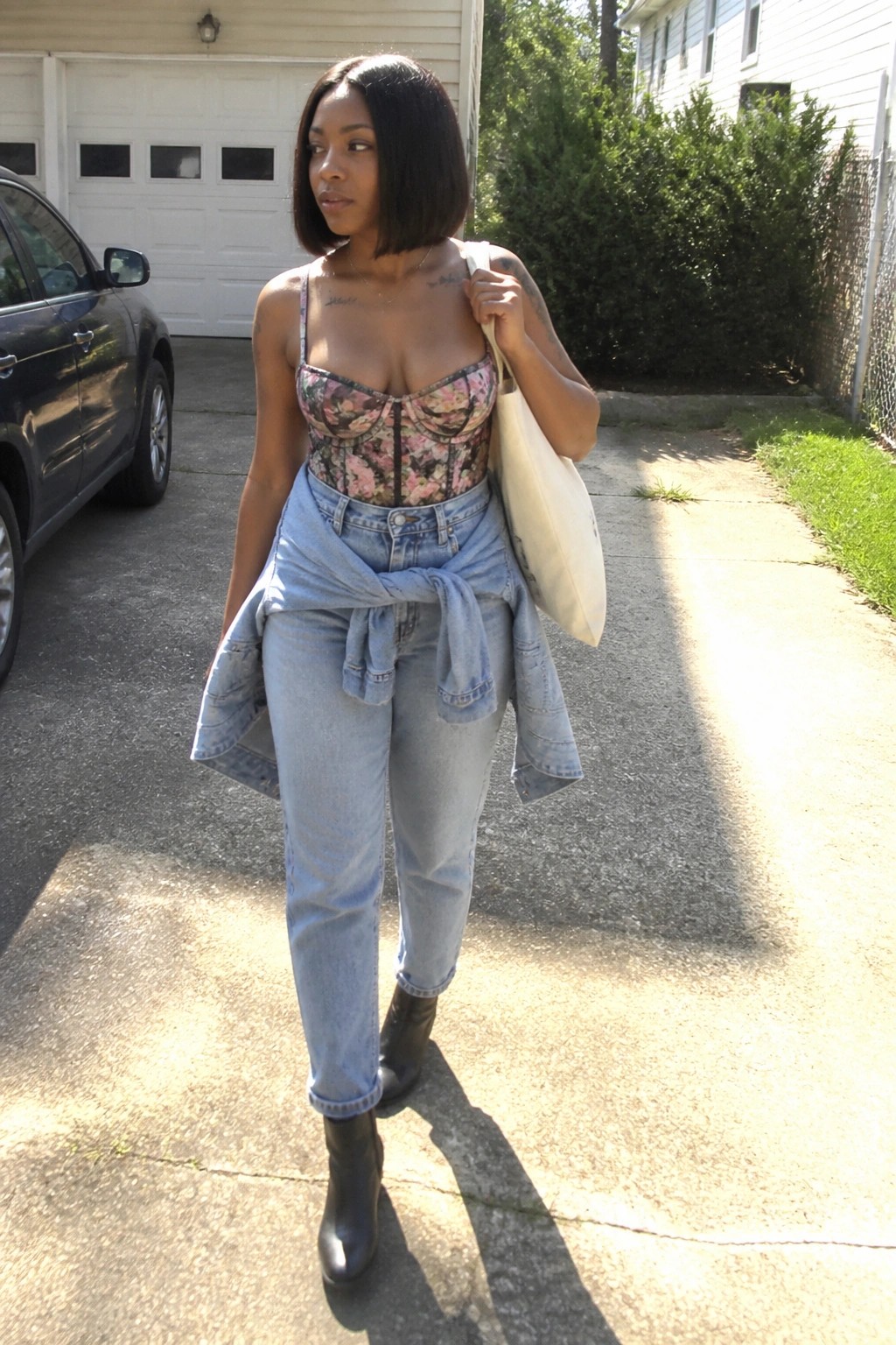 Woman in floral corset top and jeans with tied jacket holding tote bag on driveway