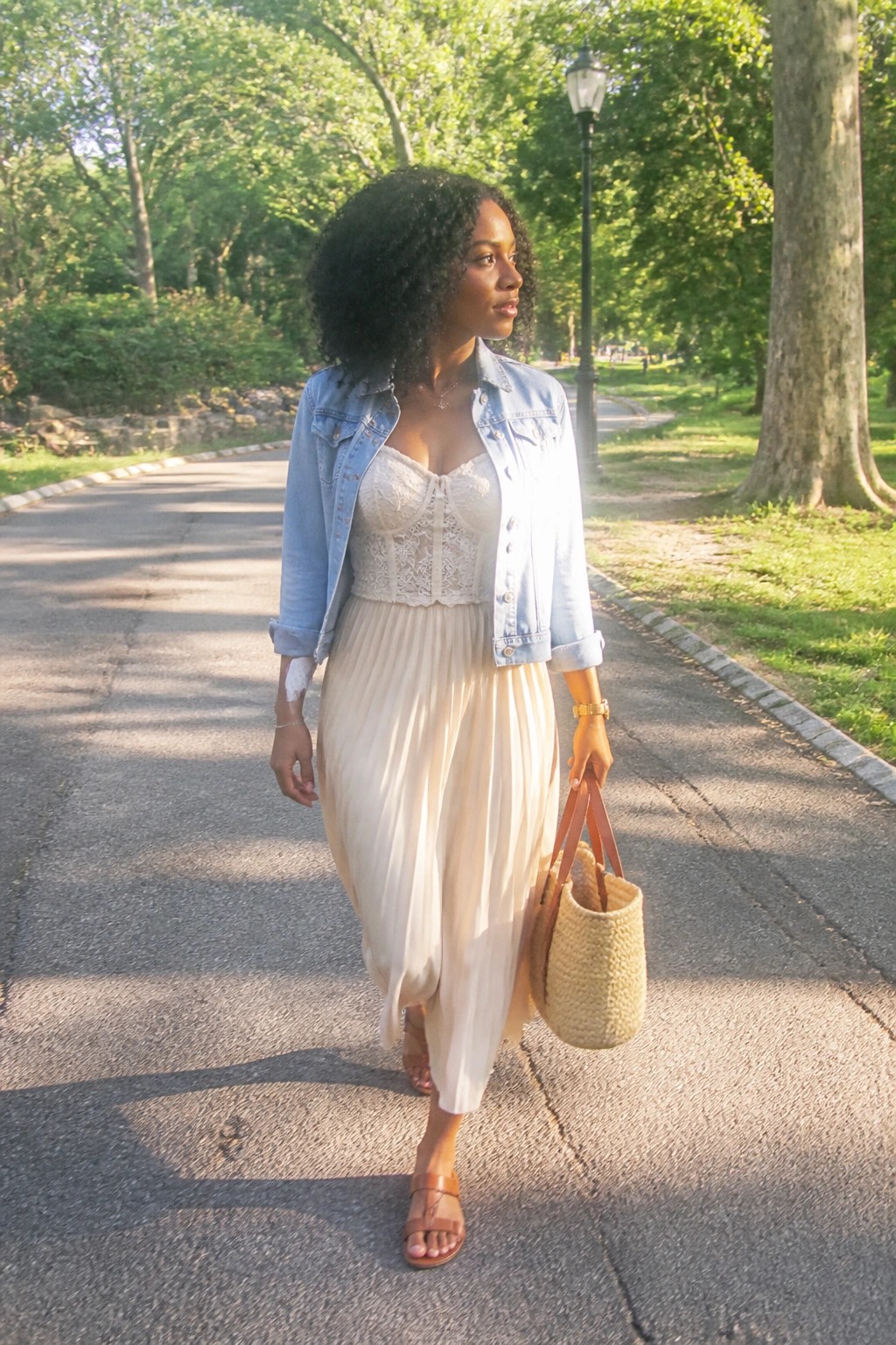 Woman in denim jacket and flowing skirt carries woven bag along sunny park path