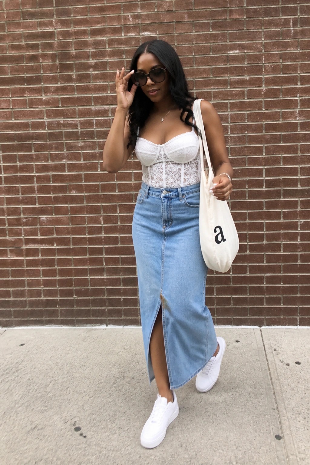 Woman in white lace corset and blue denim skirt holding tote by brick wall.