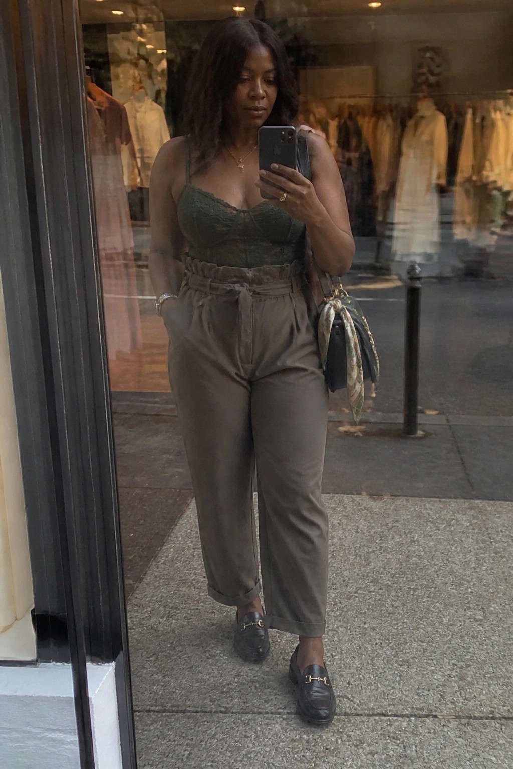 Woman in green lace top and olive belted pants taking selfie outside boutique.
