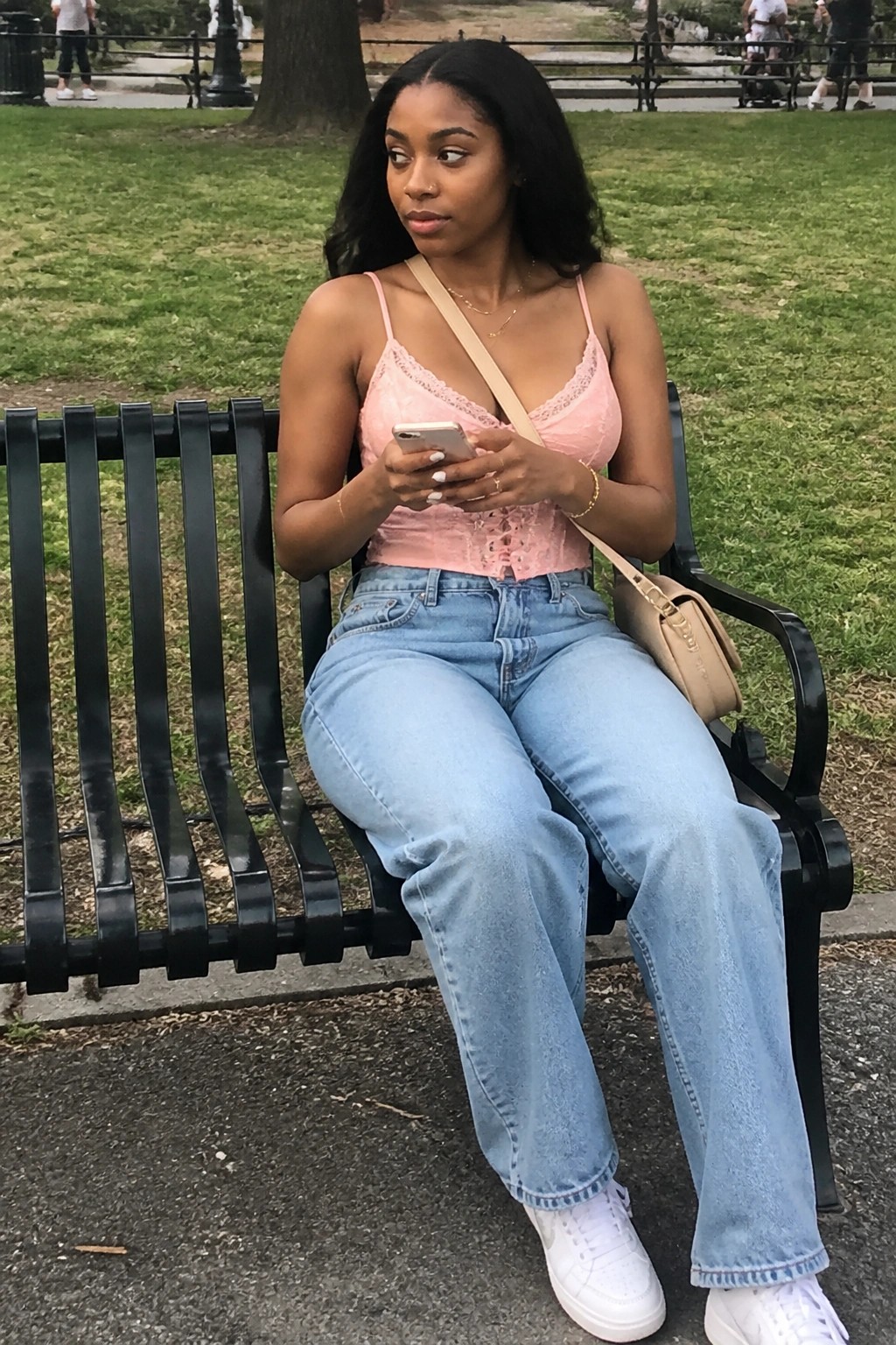 Woman in pink lace top and blue jeans sits on park bench viewing phone.